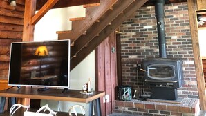Interno di una baita rustica con una scala in legno, un camino in mattoni e un televisore su un mobile in legno.