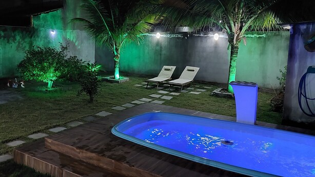 A backyard with a swimming pool, two lounge chairs, a small garden with green lighting, and a water fountain.