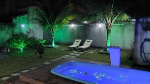 A backyard with a swimming pool, two lounge chairs, a small garden area with green lighting, and a water feature.