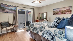 Un dormitorio con cama, una silla, un escritorio, un ventilador de techo y un puerta corrediza de vidrio que da a un balcón.