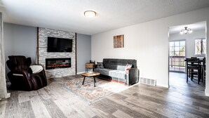A modern living room with a fireplace, a massage chair, a gray sofa, and a dining area with a table and chairs. - Big Sky Hotelier - Outstanding Sauna House (Billings)