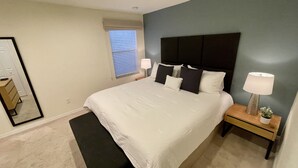 Un dormitorio con una cama grande, mesitas de noche, un banco, una ventana con persianas y un espejo.