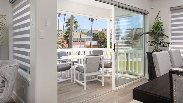 A modern outdoor patio with a white table and chairs, a glass door leading to a balcony, and a potted plant.