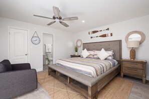 A bedroom with a bed, a bench, a nightstand, a lamp, a ceiling fan, a clock, and a door. - Two Minute Walk to Beach (South Padre Island)