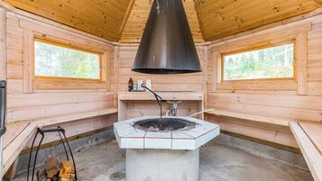 A log cabin interior with a central fireplace, wooden walls, and two windows.