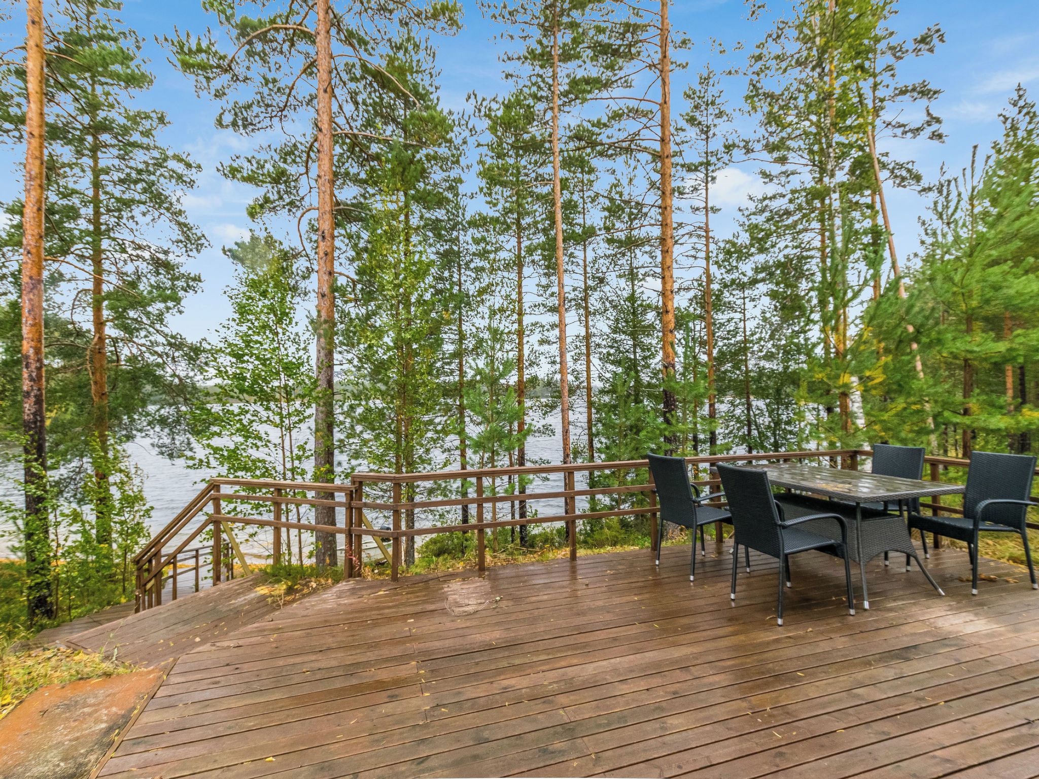 Eine Holzterrasse mit Tisch und Stühlen bietet Blick auf einen See, umgeben von hohen Bäumen.