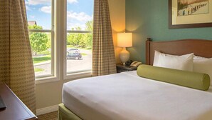 Habitación de hotel con una cama grande, una mesita de noche con lámpara, una ventana con cortinas y un cuadro en la pared.