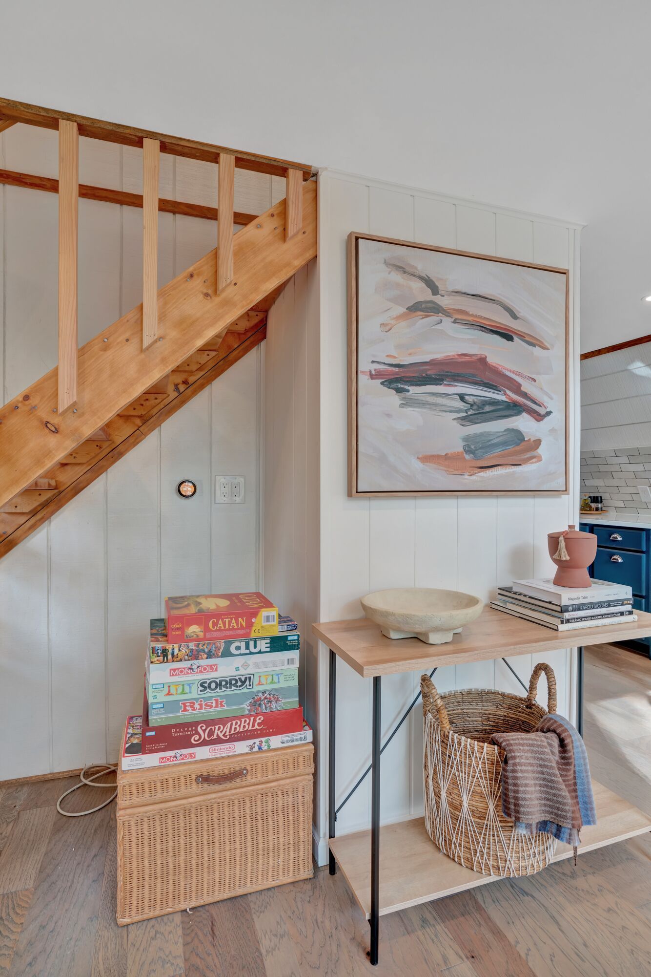 Een trap met houten leuningen, een aan de muur bevestigd abstract schilderij, een tafeltje met een schaal en boeken, en een rieten mand op de vloer.
