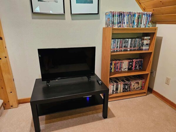 A black TV stand with a remote control, a bookshelf filled with DVDs, and framed pictures on the wall.