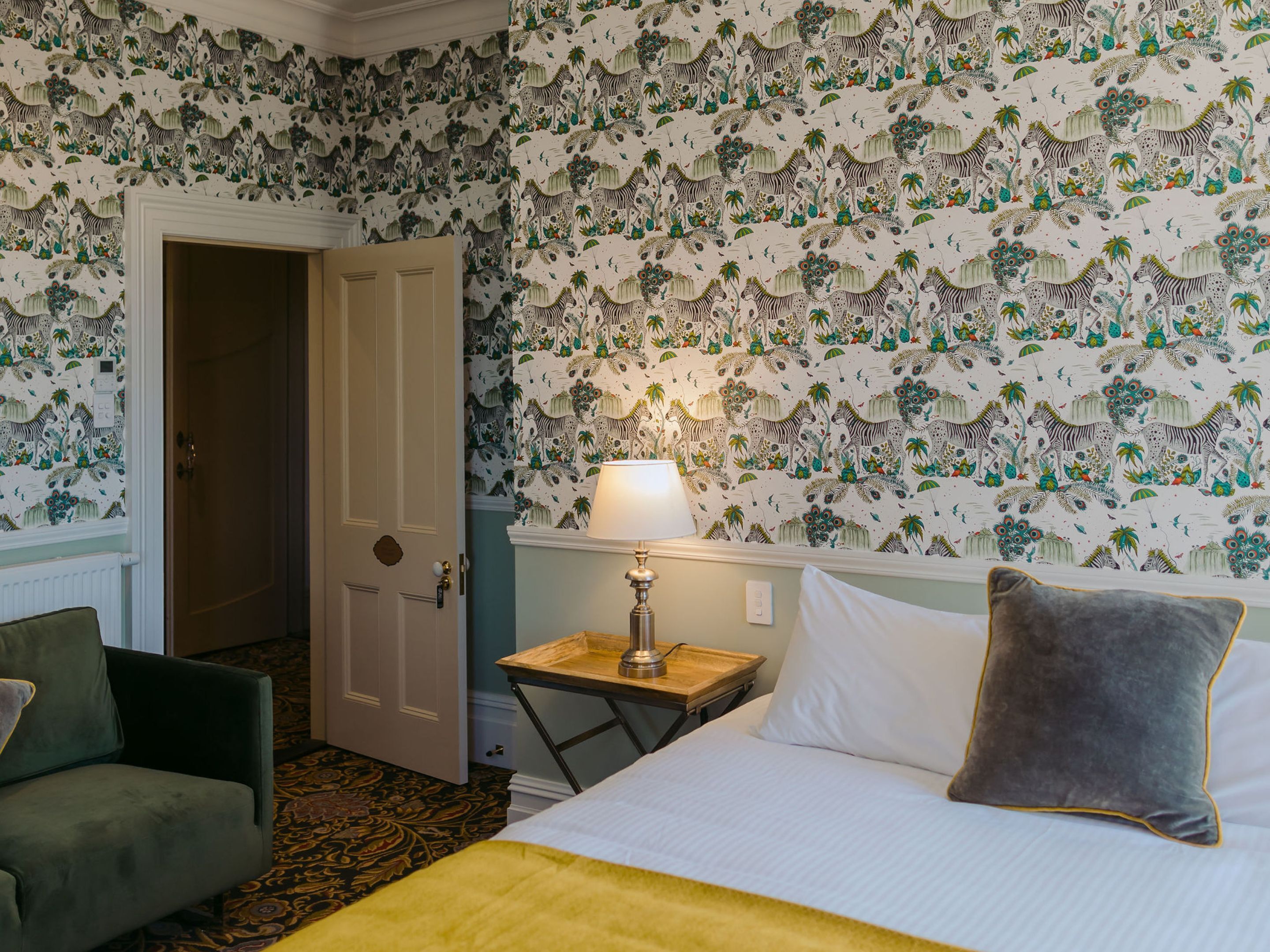 Una habitación de hotel con cama, mesita de noche, lámpara, silla y puerta.