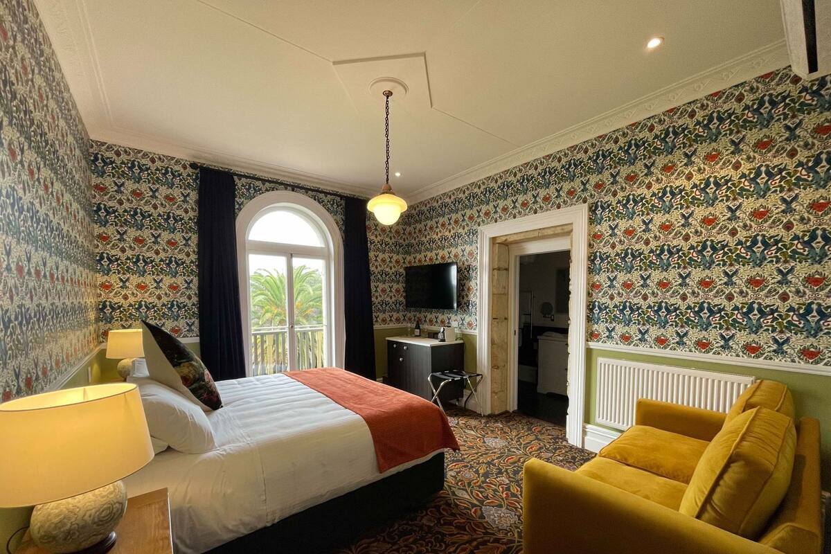 Habitación de hotel con cama, escritorio con lámpara, televisión y un sillón amarillo, además de una alfombra estampada.