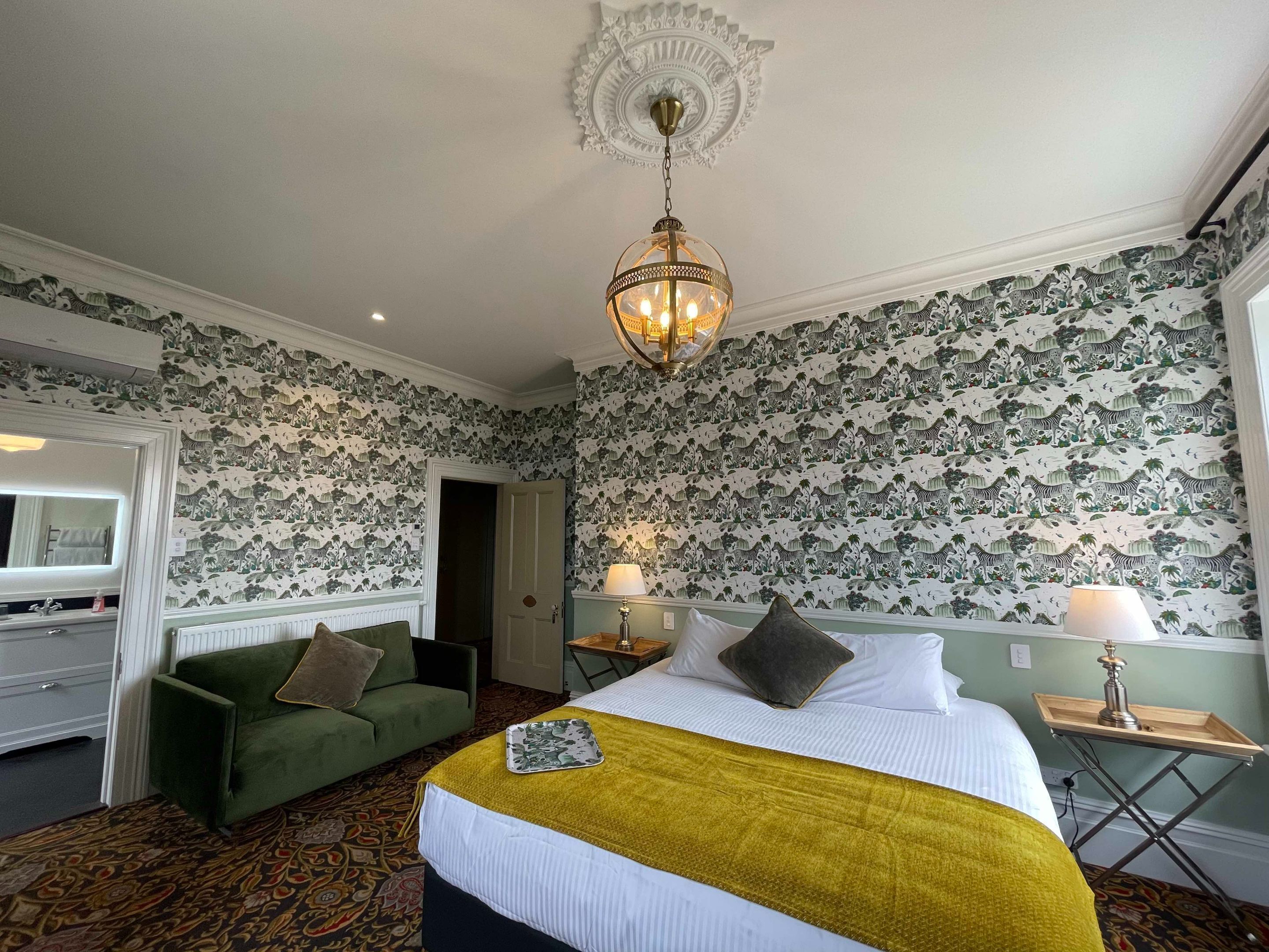 A hotel room with a bed, green sofa, patterned wallpaper, and a chandelier.