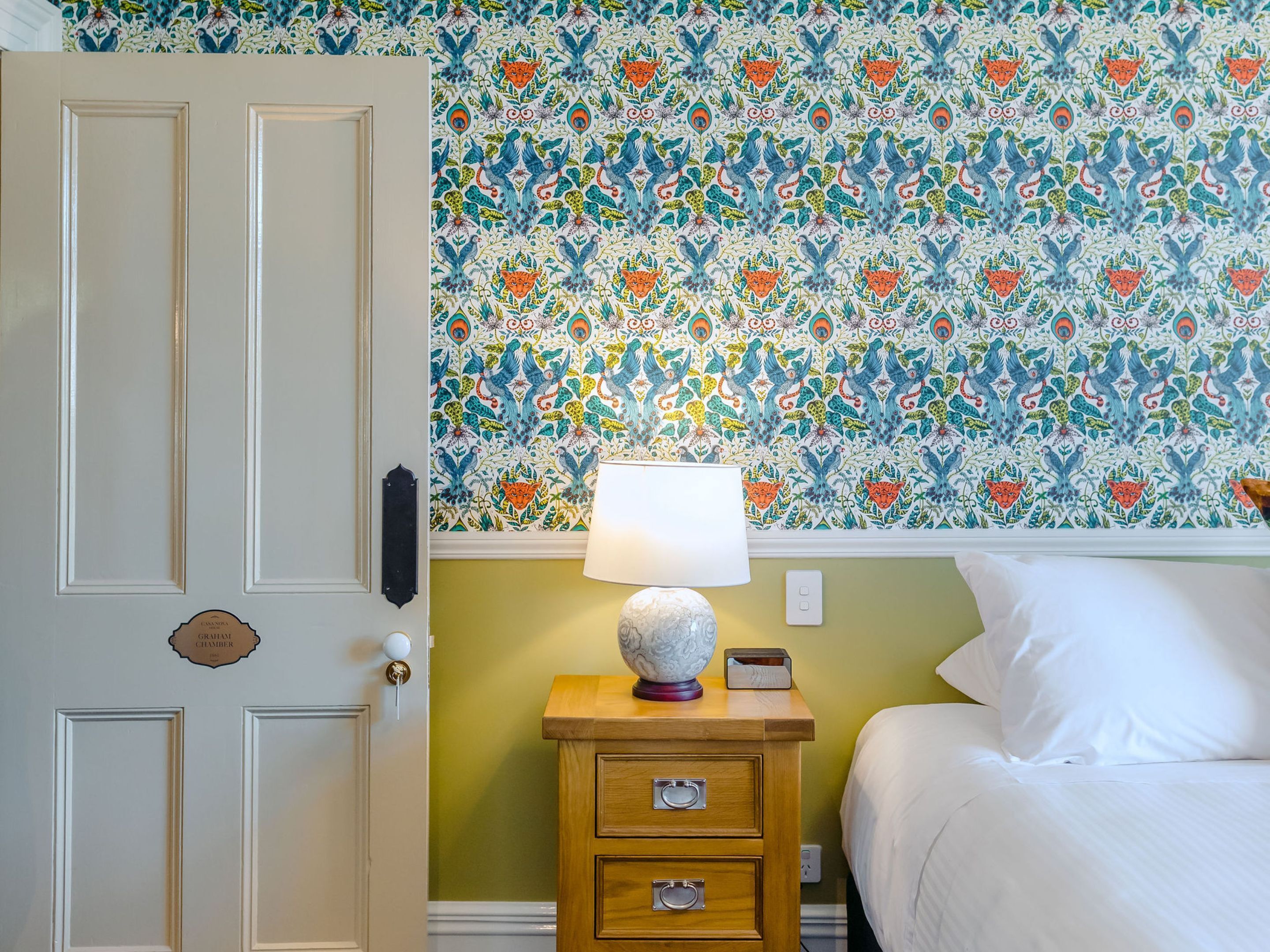 Habitación de hotel con papel tapiz estampado, una mesita de noche de madera con cajones, una lámpara y una cama con ropa blanca.