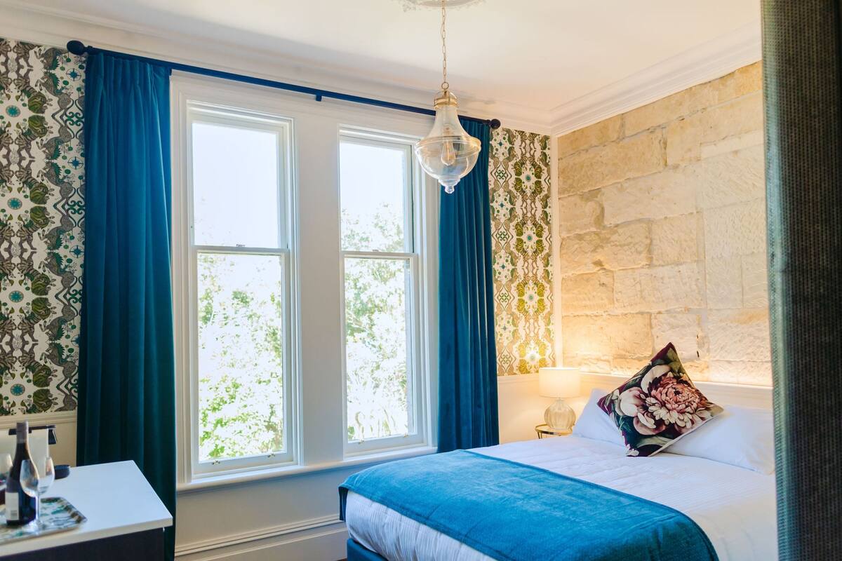Un dormitorio con dos ventanas, una cama con colcha azul, una mesita de noche con lámpara y un papel tapiz estampado.