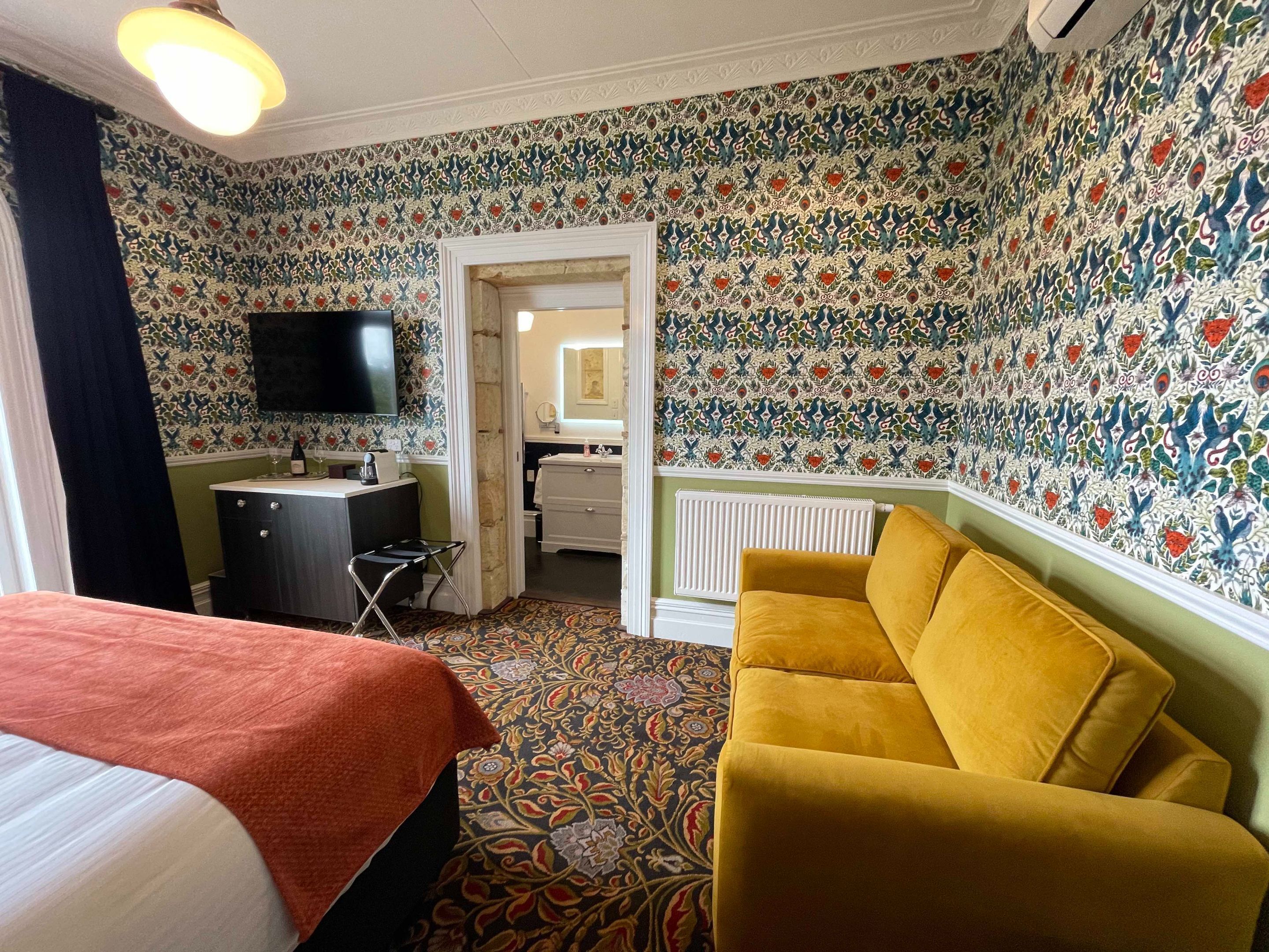 A hotel room with a bed, a yellow armchair, a desk with a TV, and a bathroom visible through an open door.