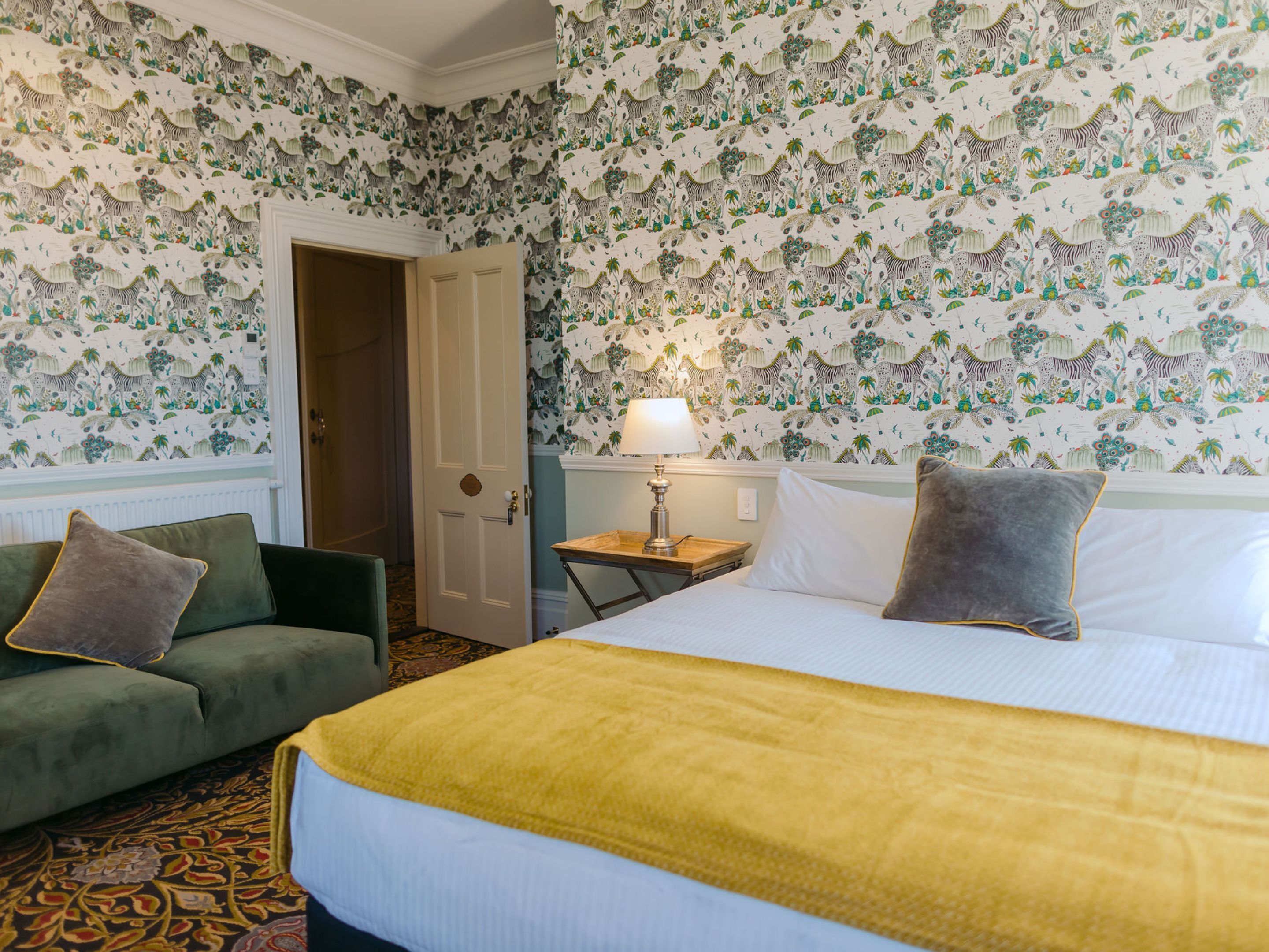 Een hotelkamer met een bed, twee fauteuils, een nachtkastje met een lamp en een deur.