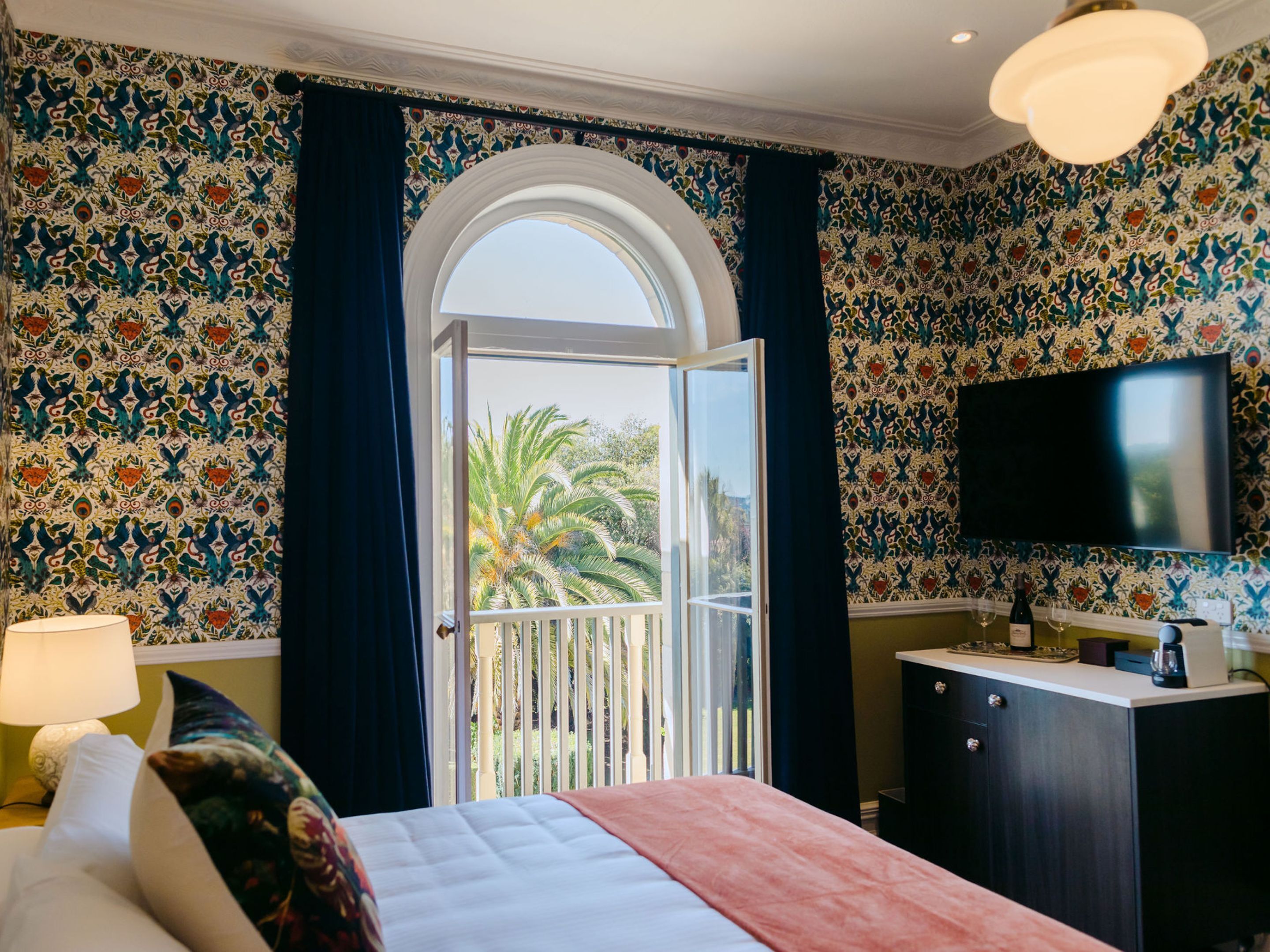 A hotel room with a bed, a desk with a TV, a window with curtains, and a balcony with a view of palm trees.