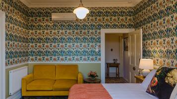 A room with patterned wallpaper, a yellow sofa, a bed with a red throw, and a hanging light fixture.