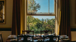 Uma sala de jantar com uma mesa posta para quatro pessoas, uma janela com cortinas e vista para árvores e um edifício.