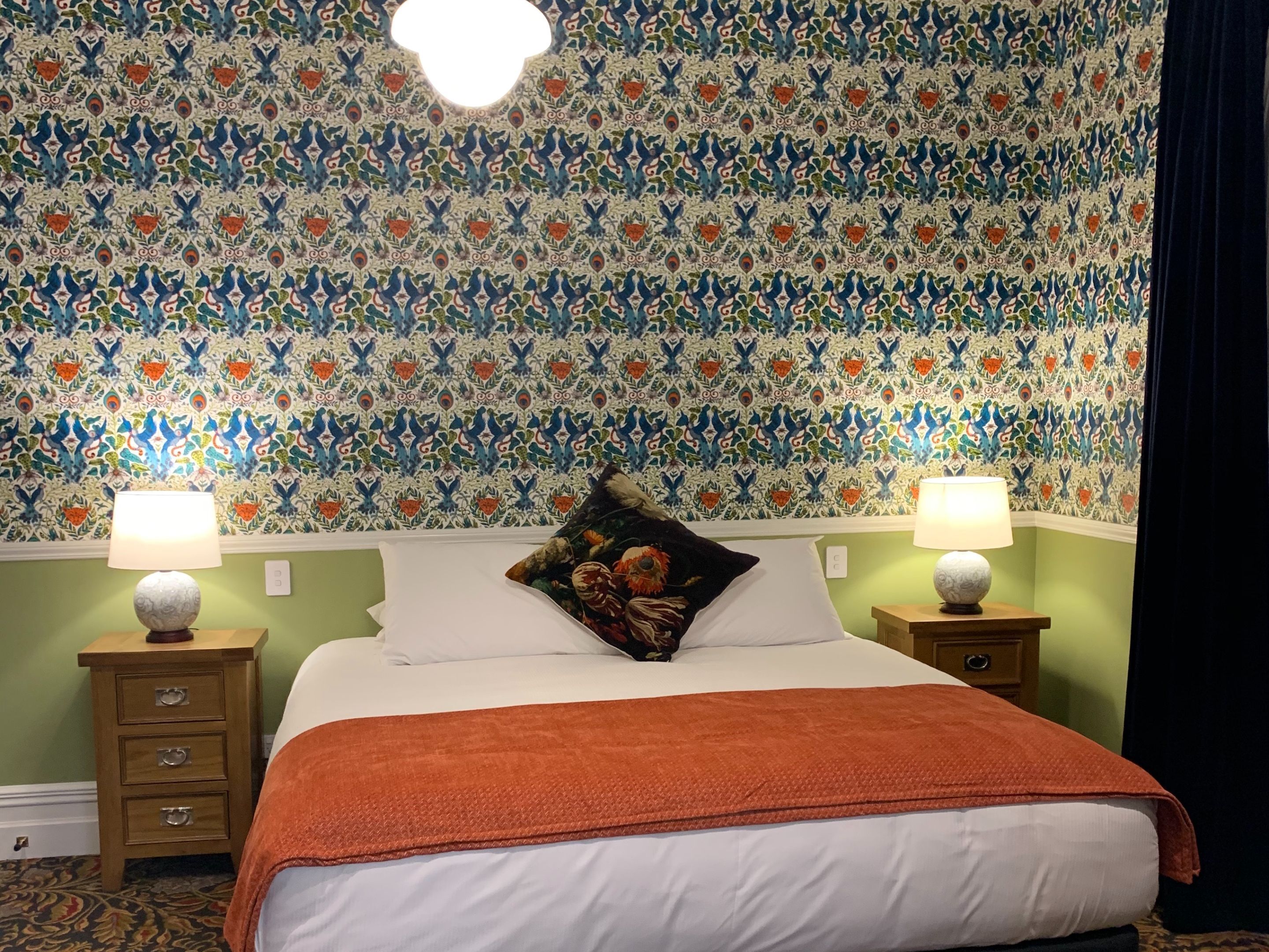 Habitación de hotel con papel tapiz estampado, dos lámparas de noche, cama con ropa blanca y una manta roja.