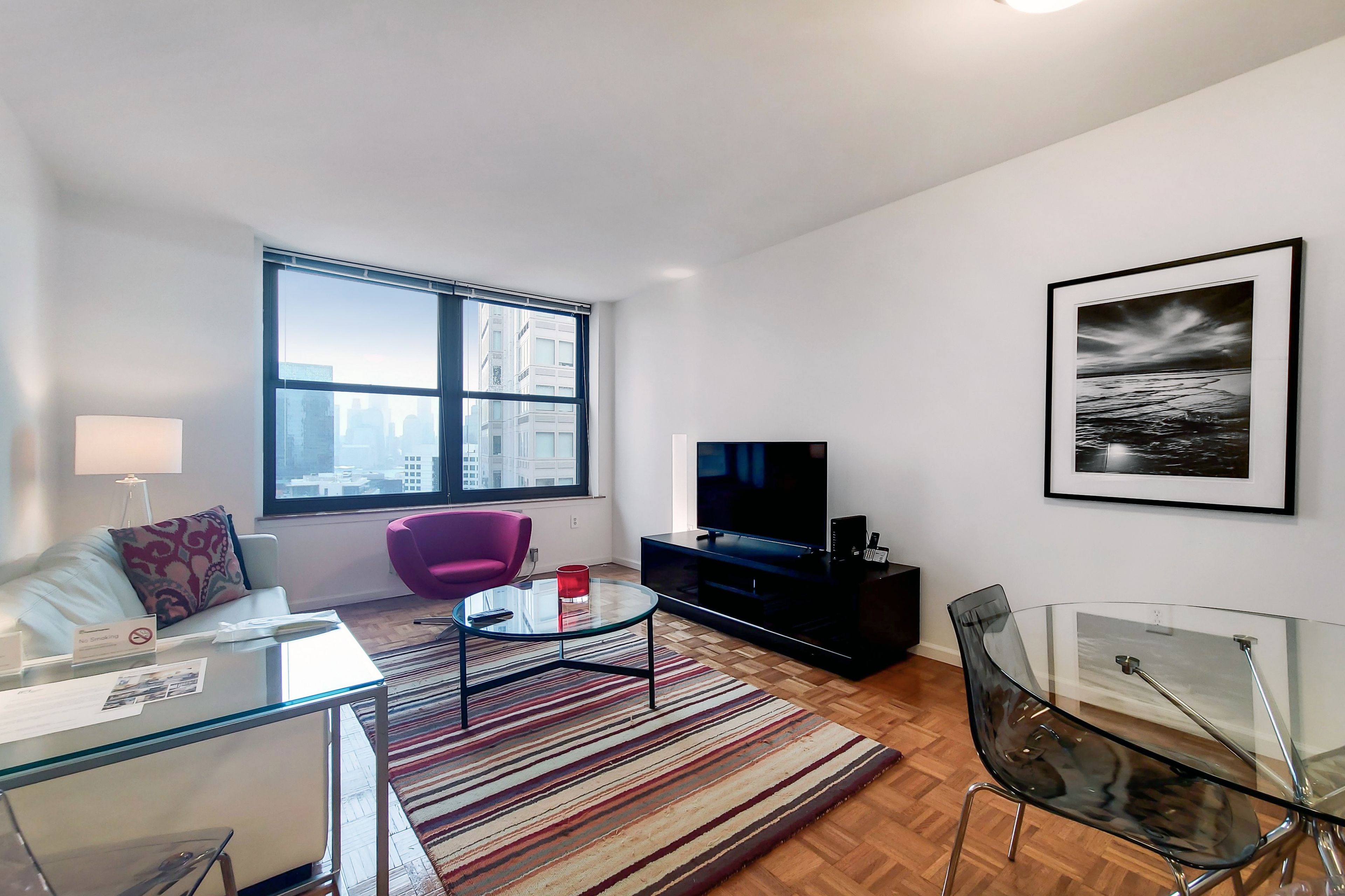 Un salon moderne avec un canapé, une table basse en verre, un fauteuil violet, un téléviseur et un tableau encadré accroché au mur.