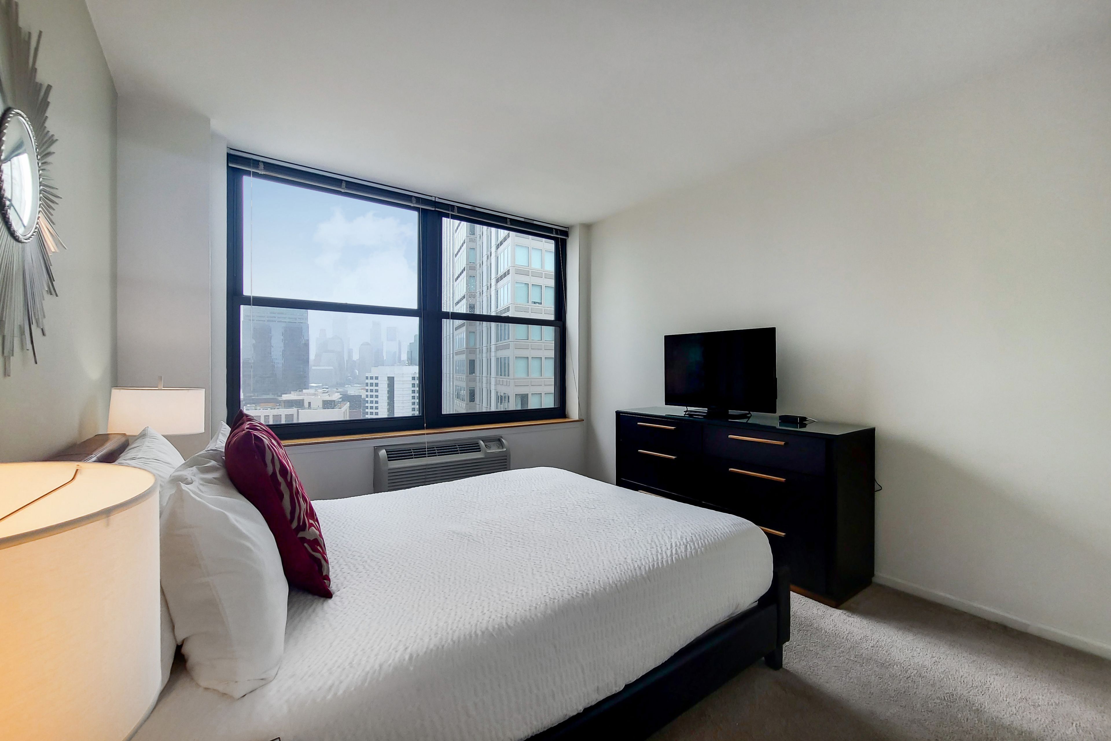 A bedroom with a large window, a bed, a TV, a dresser, and a lamp.