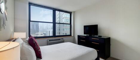 A bedroom with a large window, a bed, a TV, a dresser, and a lamp.