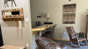 A cozy living room with a brown leather sofa, a wooden coffee table, a small wooden bench, a dining area with a wooden table and chairs, and a wall-mounted mirror.