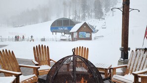 A snow-covered outdoor seating area with a fire pit, wooden chairs, and a view of a ski lift and lodge in the background.