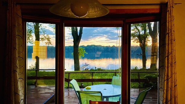 Une pièce dotée d’une grande fenêtre donnant sur un lac, d’une terrasse en bois et d’une table avec des chaises.