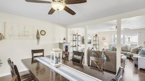 A dining area with a wooden table and chairs, a ceiling fan, and a view into a living room with a couch and a TV.