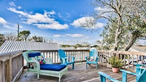 A wooden deck with wicker chairs and a lounge chair, surrounded by trees and a clear sky.