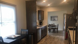 Una cocina moderna con armarios oscuros, una zona de comedor con mesa y sillas, y vista del salón a través de un arco abierto.