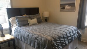 Una cama bien hecha con un edredón gris y blanco a rayas, una mesita de noche con una lámpara y un cuadro en la pared.