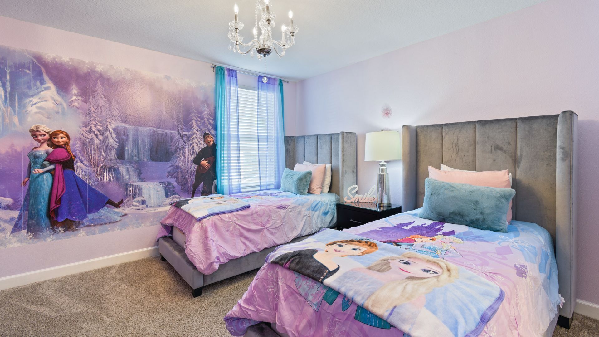 A bedroom with two beds, a mural of Frozen characters, a chandelier, and a window with blue curtains.
