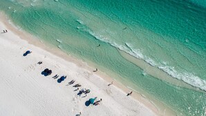 Vue aérienne d’une plage avec une eau turquoise cristalline, du sable blanc, ainsi que plusieurs parasols et chaises longues.