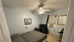 Un dormitorio con cama, una silla, ventilador de techo, un cuadro enmarcado, un espejo y un piano.