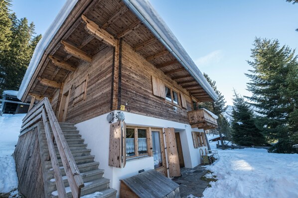 A wooden cabin with a staircase, a covered porch, and a snow-covered ground.