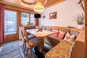 Interior acolhedor de uma cabana com uma mesa de jantar de madeira, sofá estampado e uma televisão na parede.