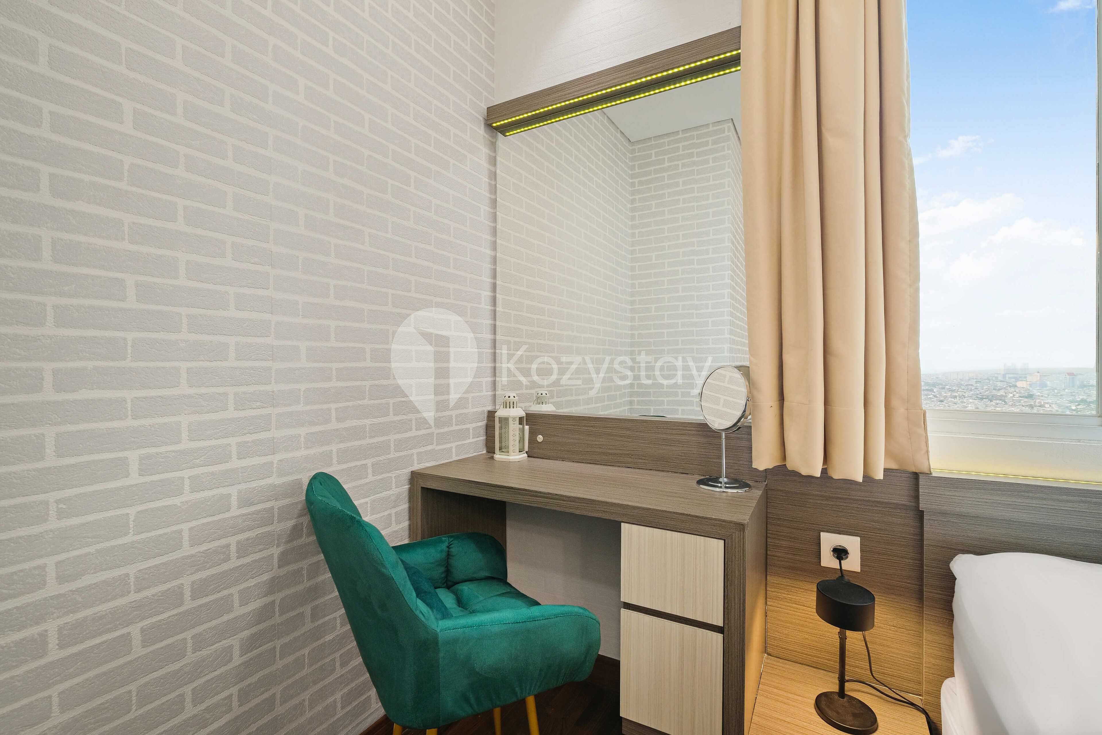 A modern hotel room with a brick-patterned wall, a desk with a mirror, a teal chair, a bedside table with a lamp, and a window with curtains.