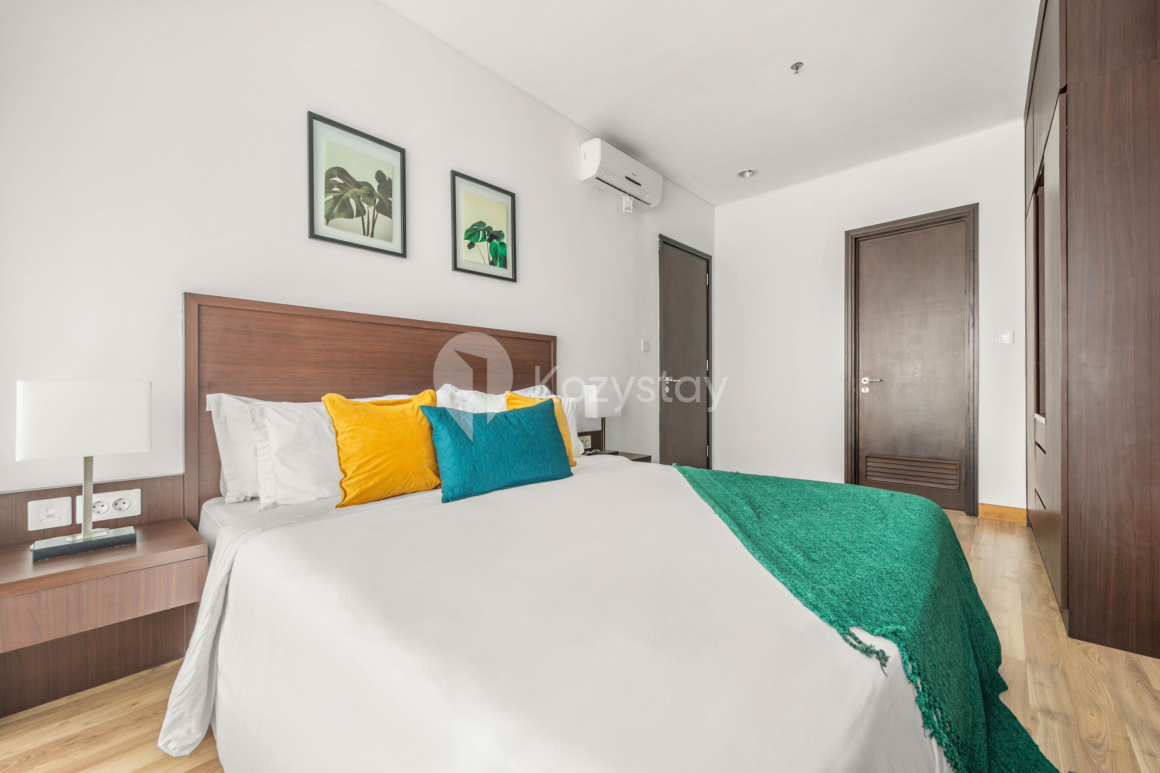 A modern hotel room with a large bed, a bedside table, a nightstand, a wall-mounted air conditioner, and two framed pictures.