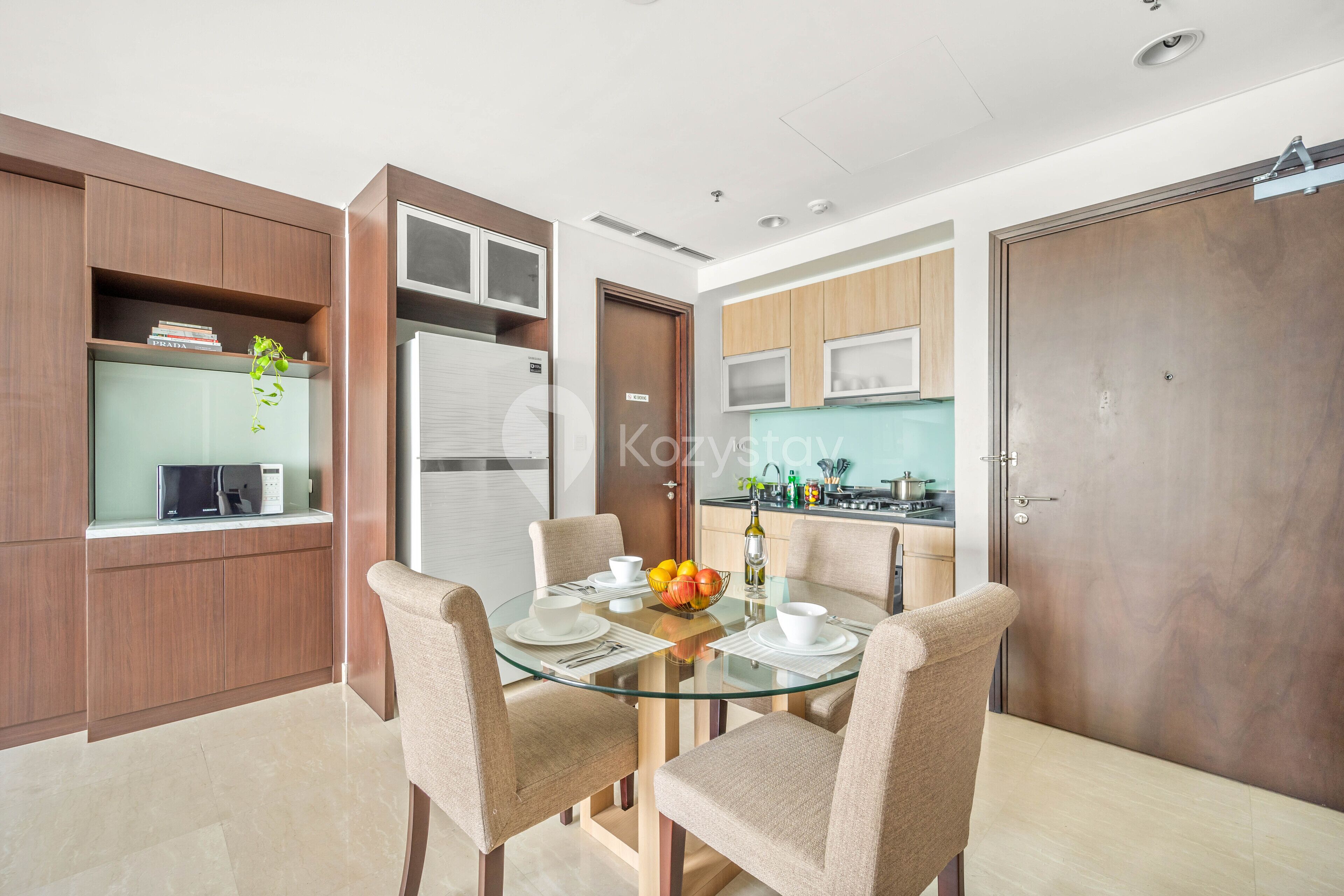 A modern kitchen with a microwave, refrigerator, and wooden cabinets. There is a dining area with a glass table and beige chairs.