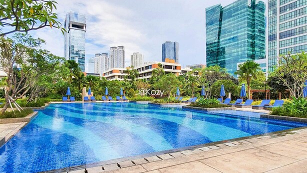 A swimming pool with lounge chairs and umbrellas, surrounded by greenery and modern buildings in the background. - LONDON by Kozystay (Jakarta)