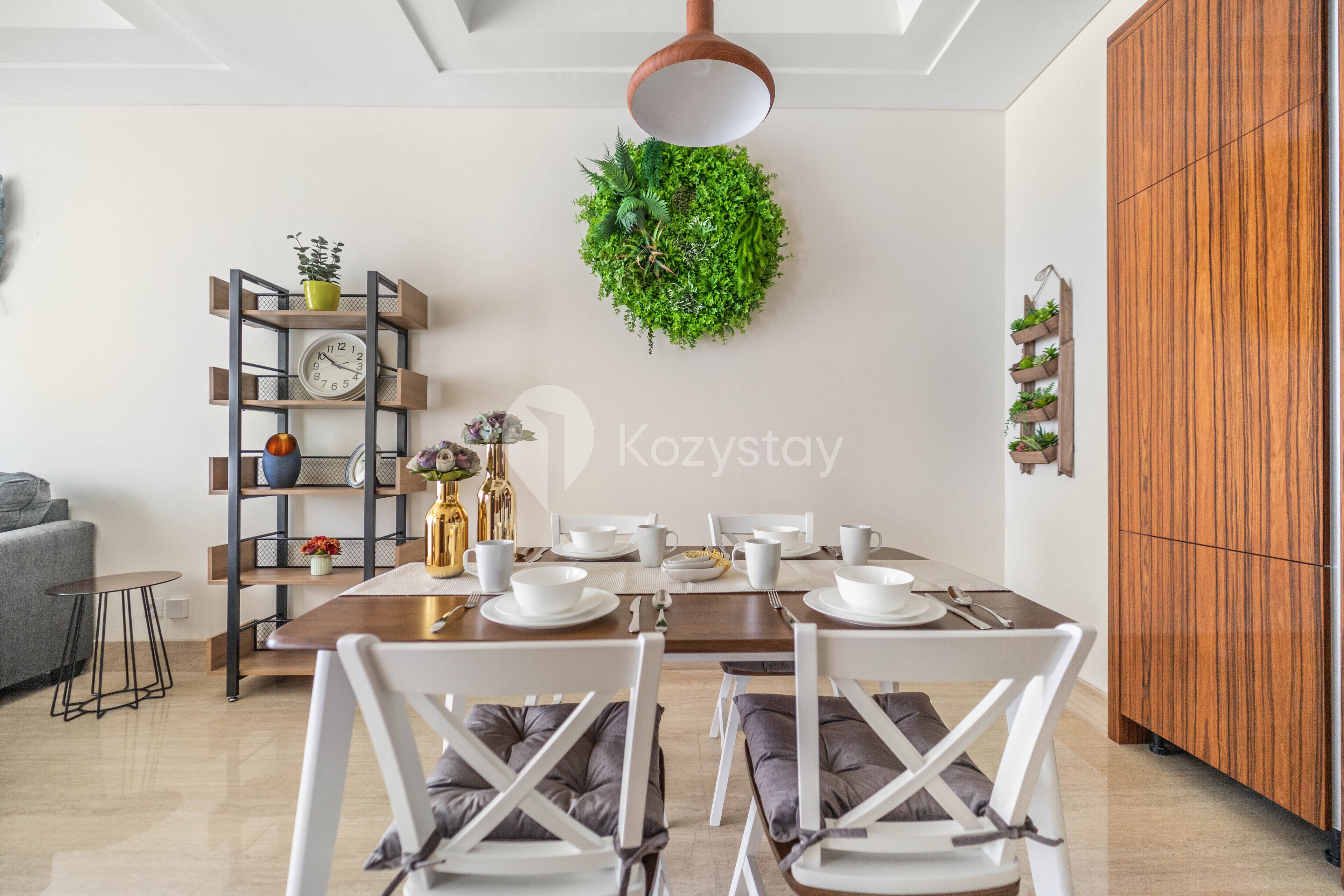 Un coin repas avec une table en bois dressée pour quatre personnes, entourée de chaises blanches avec des coussins gris. Une plante verte suspendue se trouve au-dessus de la table, et un meuble en bois est placé sur la droite.
