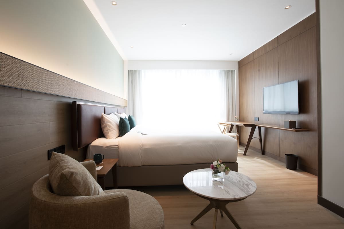 A modern hotel room with a large bed, a flat-screen TV, a round coffee table, and a seating area with a sofa and armchair.