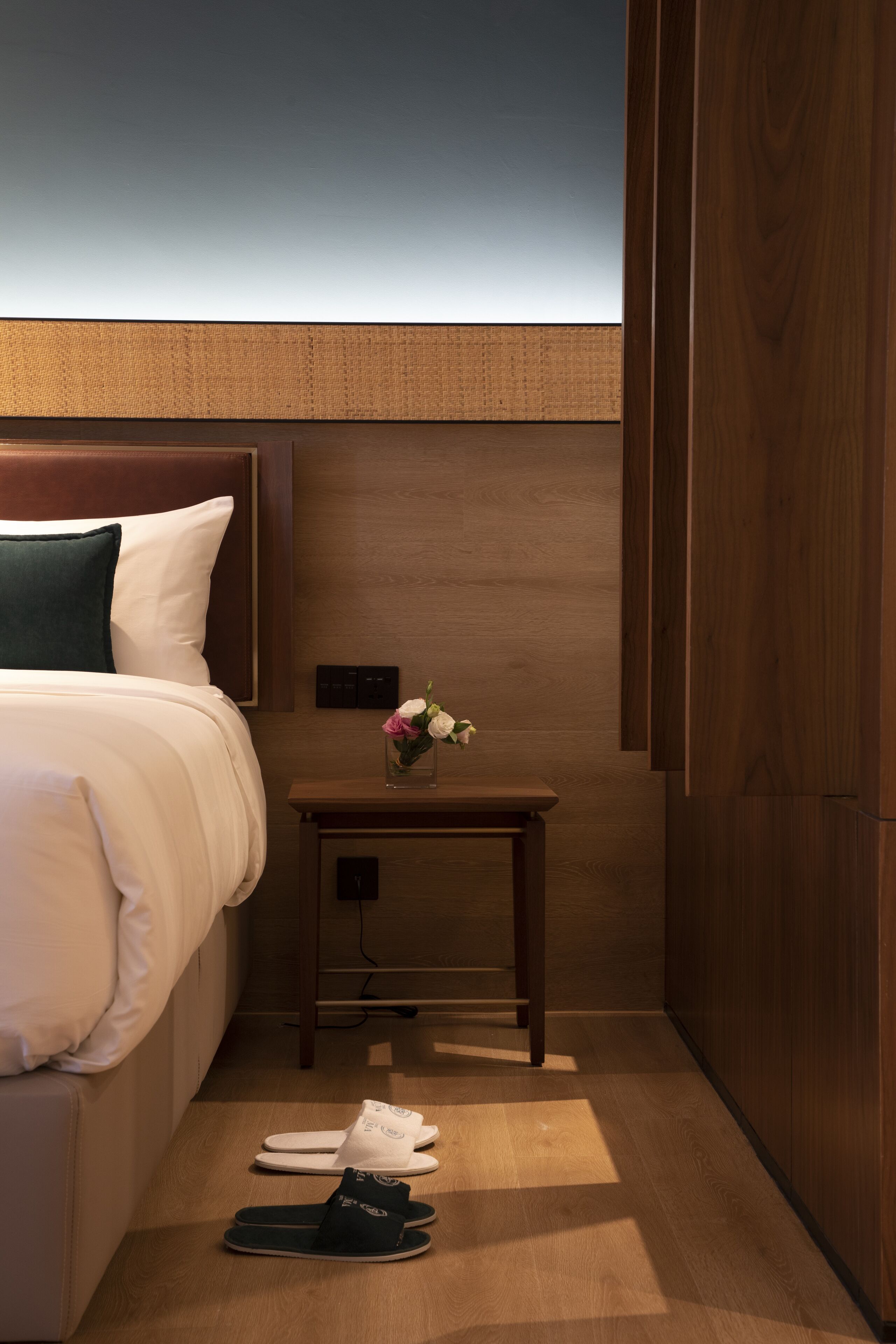 A hotel room with a bed, a nightstand with a vase of flowers, and a pair of slippers on the floor.