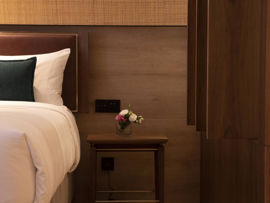 A hotel room with a bed, a nightstand with a vase of flowers, and a pair of slippers on the floor.