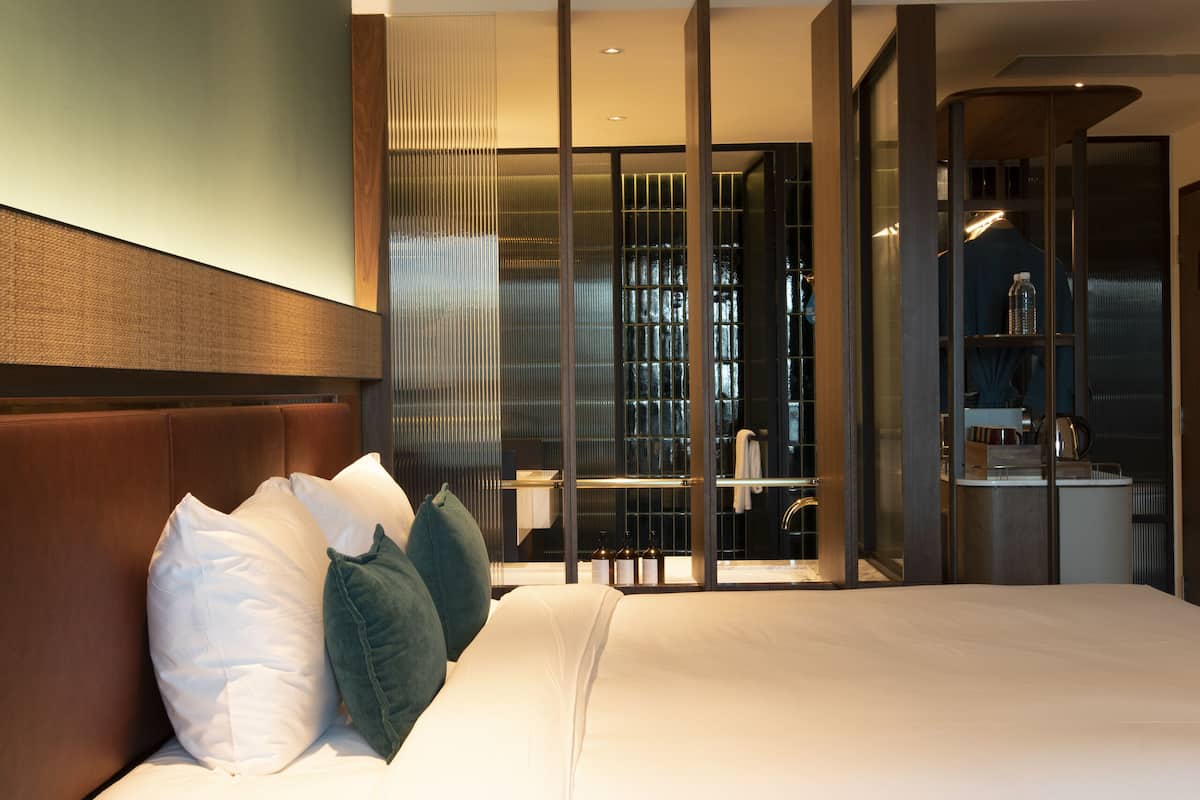 A modern hotel room with a large bed, a nightstand, and a bathroom visible through a glass partition.