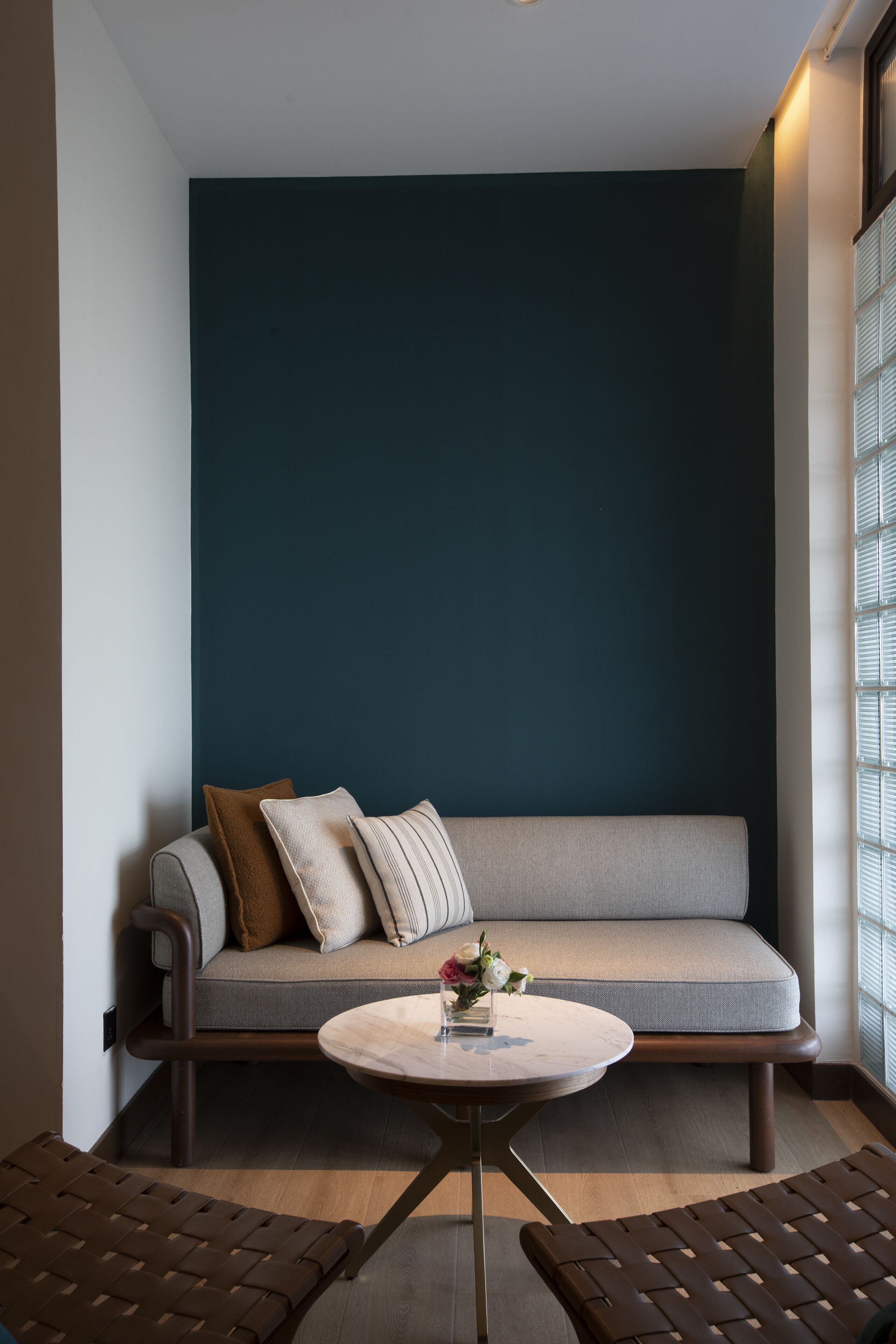 A corner room with a dark teal accent wall, a grey sofa with cushions, a small round table with a vase of flowers, and two brown woven chairs.