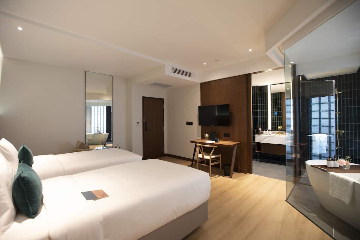 A hotel room with two beds, a desk, a TV, a bathroom with a mirror, and a shower.